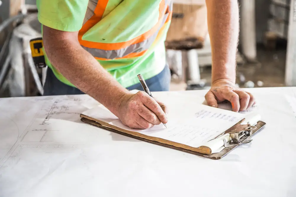 A worker in a high-visibility shirt writes on a clipboard resting on a large printed plan.