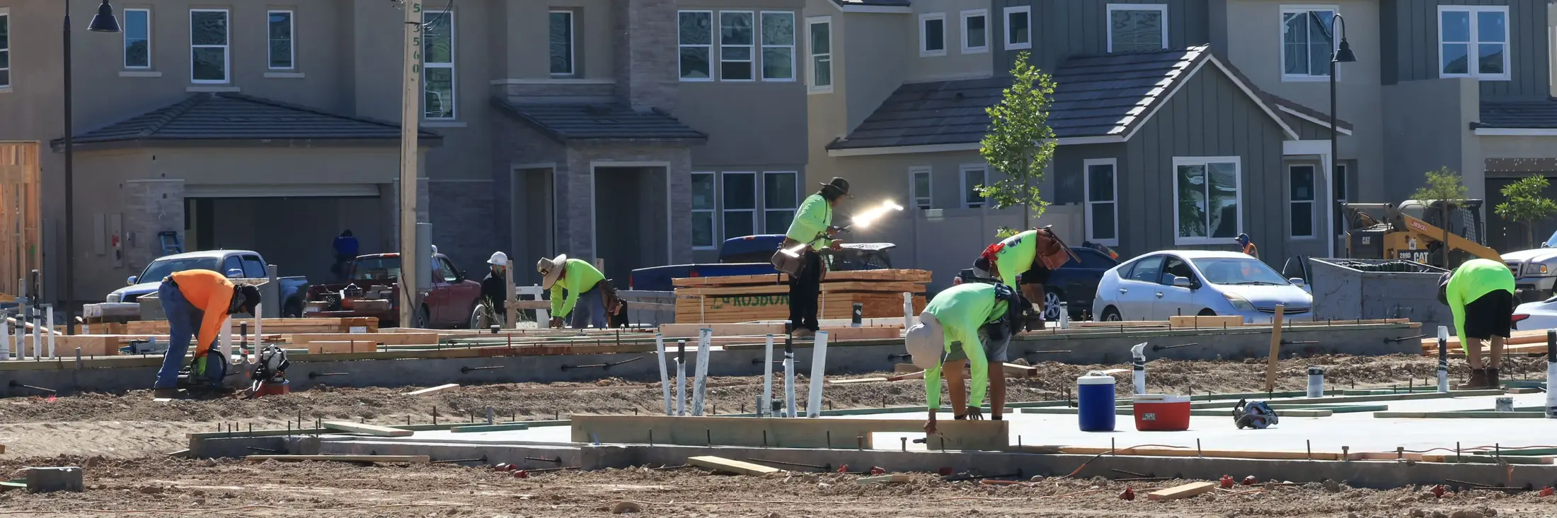 A crew in bright shirts works on new foundations in a subdivision, with finished houses and parked trucks behind them.