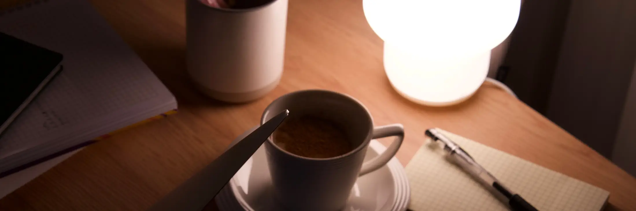 A desk at night lit by a small round lamp, with a cup of coffee, a pen cup and a notebook with a pen.