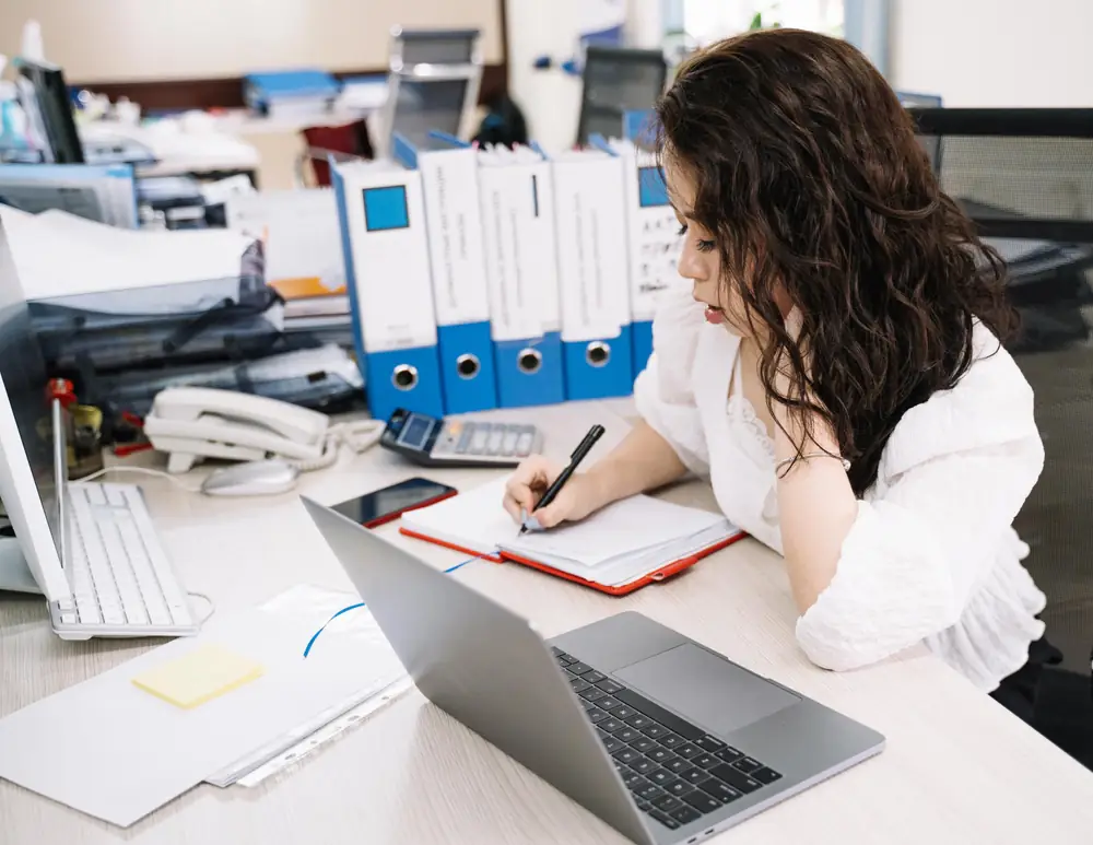 An office worker takes notes at a desk lined with binders, next to an open laptop, a desk phone and a calculator.